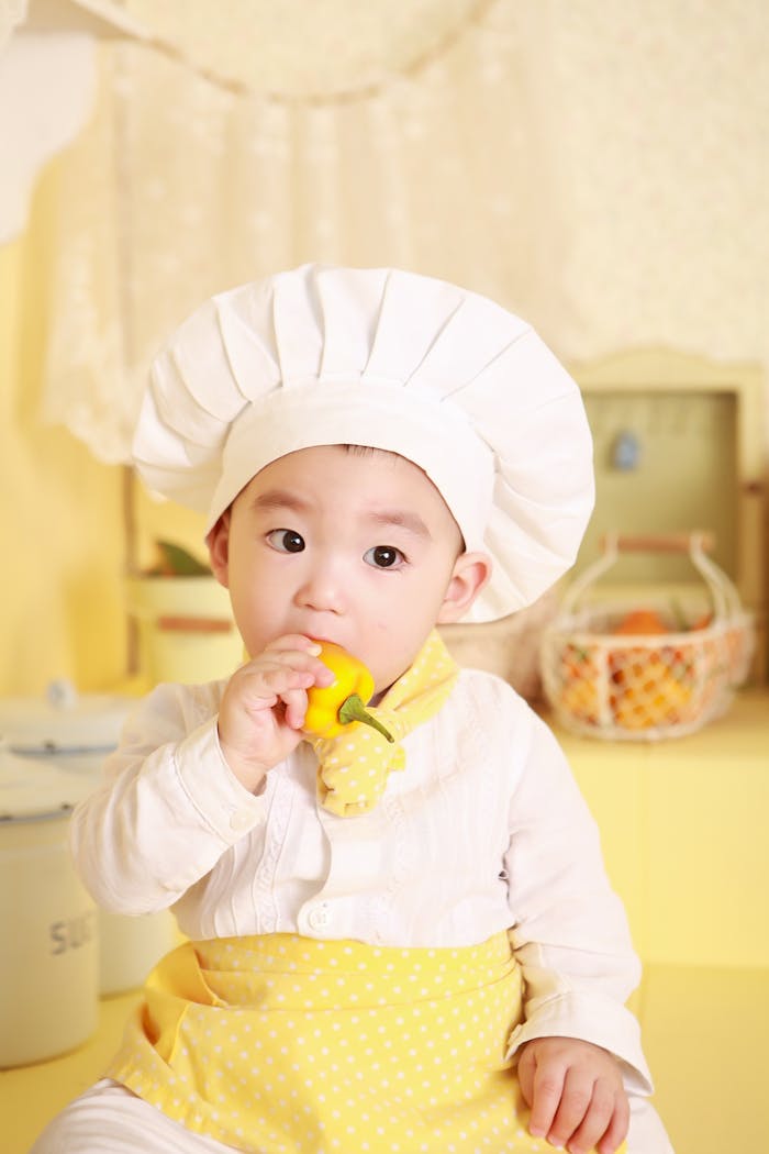 Cute baby in chef hat and apron nibbling a bell pepper in bright kitchen.