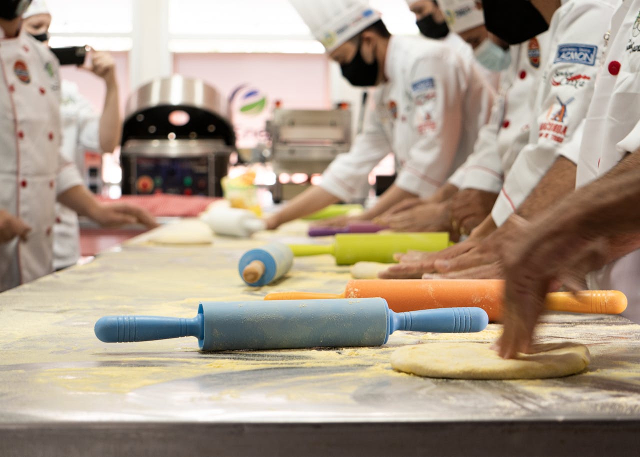 Chefs wearing uniforms and masks prepare dough with colorful rolling pins in a professional kitchen.